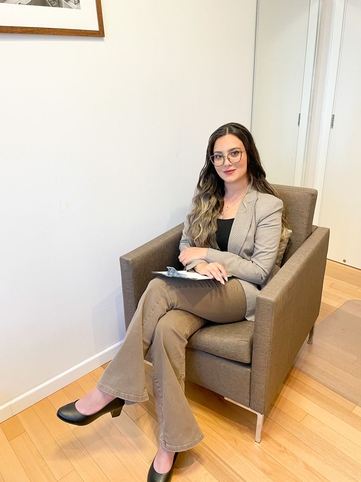 Psicóloga Juliana Paixão sentada em uma poltrona em seu consultório, vestindo blazer xadrez e calça social
